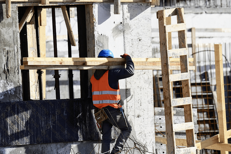 Man in a high visibility vest and hard hat carrying a plank of wood over his shoulder