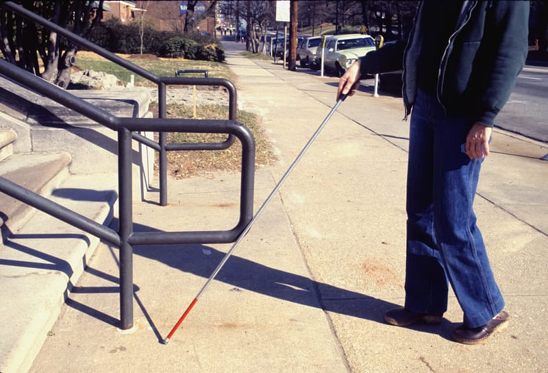 A man with a grey cane is walking up a set of stairs. He appears to be blind and is carefully feeling his way with the cane to avoid tripping or stumbling.